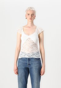 Hvit blonde-topp med korte cap-ermer, med et blomstermønster og bølgekant i halsen, kombinert med lyseblå denimjeans.