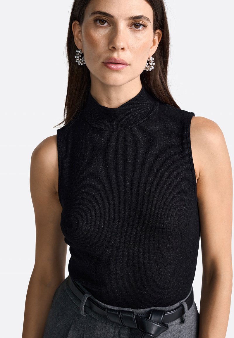 Zwart geribbelde mouwloze top met een hoge hals en een zachte textuur, gecombineerd met een zwarte riem. Model heeft zilveren studs in haar oren.