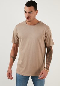 Tricou bej cu mânecă scurtă, gât la roud, textură netedă și croială lejeră. Prezintă un tatuaj detaliat pe brațul modelului.