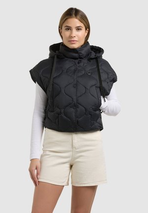 Frau trägt eine schwarze, gesteppte ärmellose Kapuzenweste über einem weißen, langärmeligen Oberteil und beigen Shorts, steht vor einem einfarbigen hellen Hintergrund.