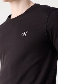Czarna koszulka z okrągłym dekoltem i małym białym haftowanym logo "cK" na lewej piersi, noszona przez osobę o jasnej karnacji.