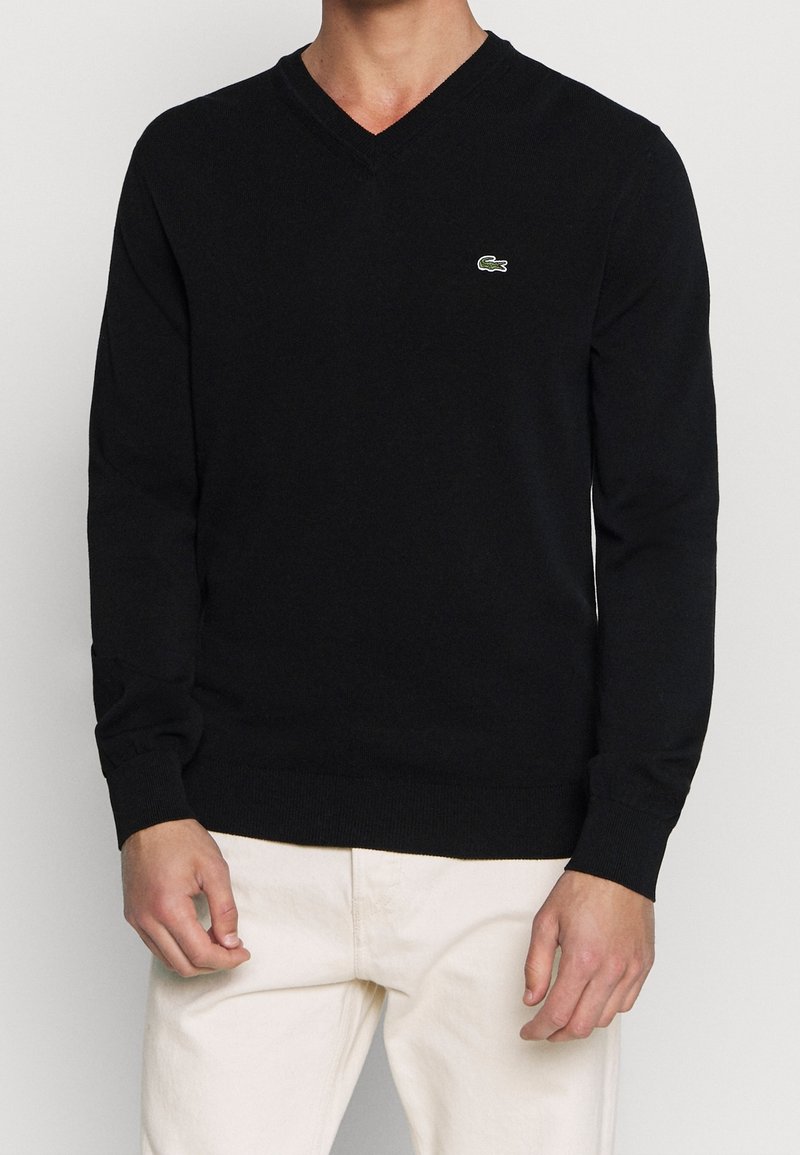 Schwarzer V-Ausschnitt-Pullover aus feinem Strickstoff mit langen Ärmeln, der ein kleines grünes Lacoste-Krokodil-Logo auf der linken Brust trägt.