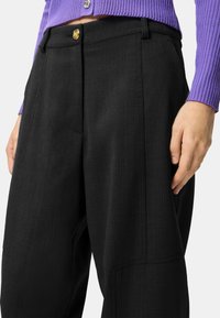 Pantaloni neri su misura con finitura strutturata, caratterizzati da una chiusura con bottone dorato e tasche laterali. Abbinati a un maglione viola a coste.