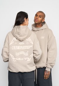 Felpa beige oversize con cappuccio regolabile, caratterizzata da testo e grafiche stampate sul retro. Realizzata in un morbido tessuto testurizzato.