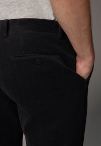 Schwarze Cord-Hosen mit strukturierter Oberfläche, ausgestattet mit einer Gesäßtasche mit Knopfverschluss und einer sichtbaren Hand in der Seitentasche.