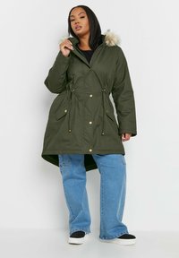 Kakifarvet parka-jacka med foder i päls, dragsko i midjan, guldtonade knappar, framfickor och längre bakställd fållen.