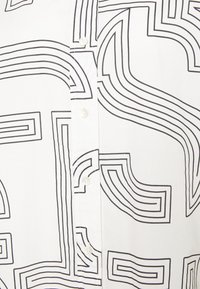 Chemise blanche avec motifs abstraits noirs en lignes, boutons visibles et texture lisse. Le motif présente des largeurs et des formes variées.