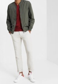 Man står med händerna i fickorna, iklädd en olivgrön bomberjacka, röd tröja, ljusbeiga byxor och vita sneakers med röda detaljer.