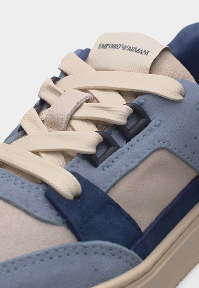 Κοντινό πλάνο ενός μπλε και μπεζ σουέντ αθλητικού παπουτσιού με μπεζ κορδόνια και λογότυπο Emporio Armani στη γλώσσα.