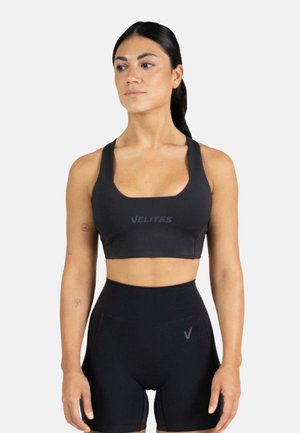 Mujer con el pelo oscuro en una coleta, vestida con un sujetador deportivo negro y pantalones cortos negros de talle alto, de pie frente a un fondo blanco liso.