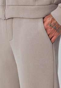 Main droite avec un tatouage floral reposant dans la poche d'un pantalon de survêtement beige, montrant un sweat à capuche beige assorti avec une fermeture éclair sur le dessus.