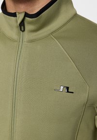 Zielona oliwkowa kurtka zapinana na zamek z czarnym kołnierzem, wykonana z gładkiego materiału, z eleganckim czarnym logo na lewej piersi.