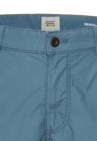 Lyseblå shorts med en tekstureret stof, der har en brun knap og stikninger. Etiketten siger "camel active" og "REGULAR."