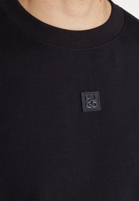Gros plan d'un t-shirt noir à col rond avec un petit patch carré portant le logo HUGO sur la poitrine.