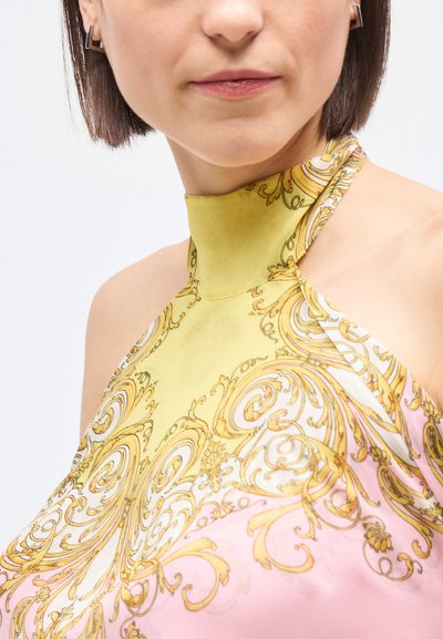 Γυναίκα με κοντά μαλλιά που φορά τοπ με ψηλό λαιμό και halter, διακοσμημένο με κίτρινο και ροζ μπαρόκ μοτίβο scroll και ασημένια τετράγωνα σκουλαρίκια.