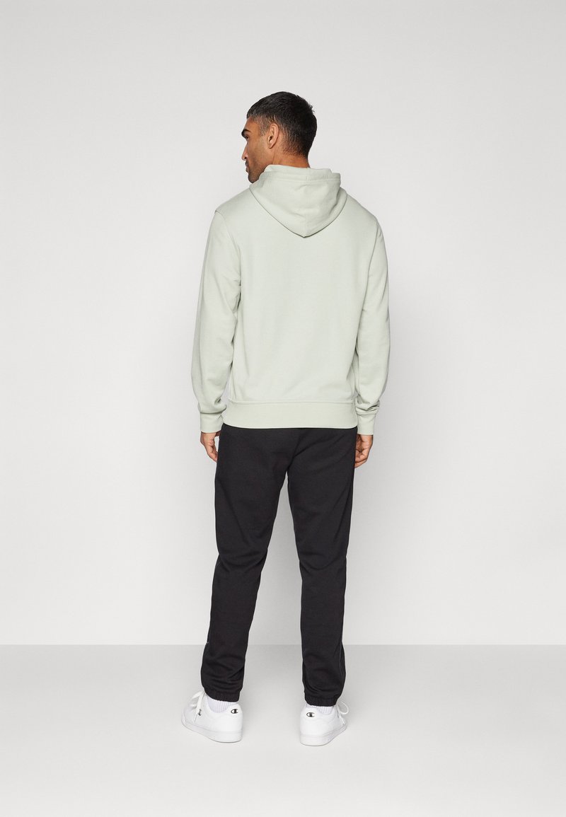Haut à capuche vert clair avec capuche et poignets côtelés, associé à un pantalon noir et des baskets blanches, vu de l'arrière sur un fond uni.