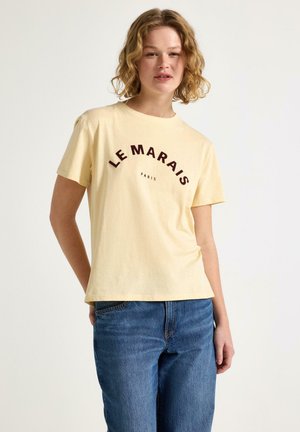 Giovane donna con capelli ricci fino alle spalle che indossa una maglietta giallo chiaro "Le Marais Paris" e jeans blu, in piedi davanti a uno sfondo semplice.
