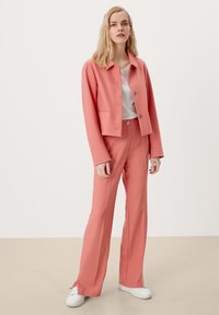 Roze op maat gemaakt pak met een cropped blazer met een knoop, bijpassende high-waisted broeken en witte sneakers op een lichtbeige vloer.