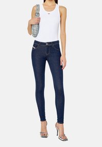 Top sin mangas blanco con un pequeño logo rojo, combinado con jeans ajustados azul oscuro y sandalias de tacón transparente. La modelo sostiene un bolso estampado.