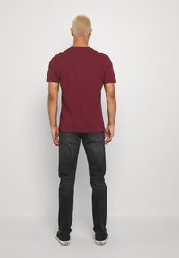 Bordowa koszulka z krótkim rękawem, zaokrąglony dekolt, noszona z ciemnymi jeansami. Luźny krój, gładka faktura, widok od tyłu.