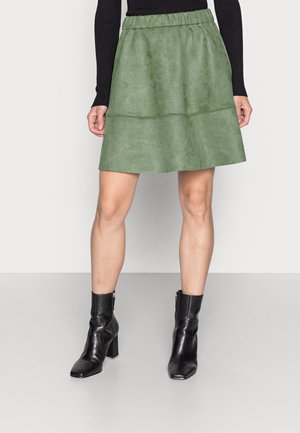 Kvinde iført en knælængde grøn ruskindnederdel med elastik i taljen, sort langærmet top og sorte ankelstøvler med hæl, stående på gråt gulv.