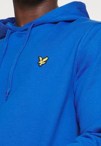 Felpa blu con cappuccio e lacci, caratterizzata da un piccolo logo ricamato di un uccello giallo sul petto, indossata da una persona con pelle scura.
