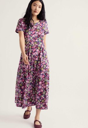 Femme portant une robe à manches courtes, longue jusqu’aux chevilles, avec un motif floral rose vif, violet et vert, et des chaussures tressées foncées, debout face à l’avant.