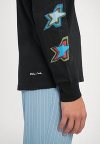 Fekete, hosszú ujjú póló, melynek ujjain színes csillaggrafikák találhatók. Texturált anyag, alján "PS Paul Smith" felirattal ellátott címkével. Kék csíkos nadrág látható.