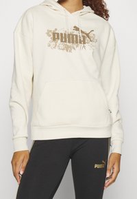 Krämfärgad huvtröja med en känguruficka och huva, med en guldig blommig Puma-logotyp. Parat med svarta leggings med guldiga accenter.