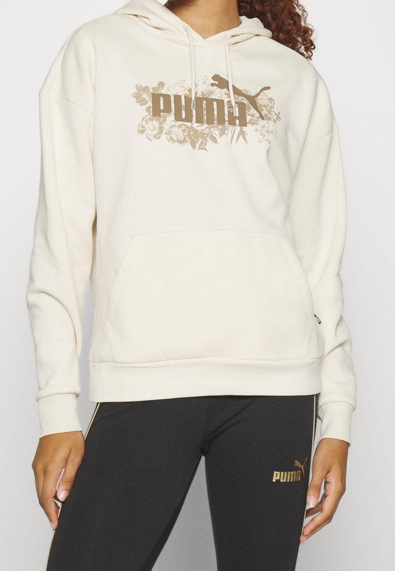 Krämfärgad huvtröja med en känguruficka och huva, med en guldig blommig Puma-logotyp. Parat med svarta leggings med guldiga accenter.