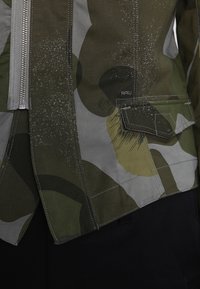 Maskovací bunda v zelených a šedých tónech, která má zapínání na zip, velké kapsy a strukturovaný povrch s různými vzory.