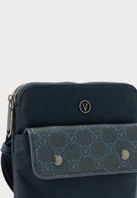 Torba tamnoplave boje s prijelaznim remenom, zatvaranjem na patentni zatvarač, prednjim džepom s preklopom koji ima uzorak na tkanini i dva metalna gumba te okruglim metalnim logotipom.