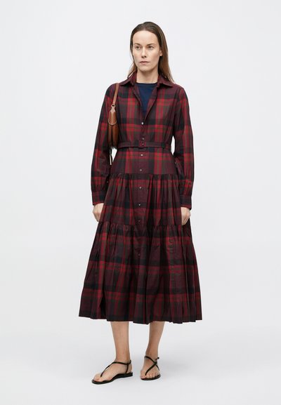 Krekla tipa kleita tumši sarkanā un melnā krāsā, ar garām piedurknēm, pogām priekšpusē, jostu viduklī un daudzslāņu svārkiem. Valkāta kopā ar melnām sandālēm.