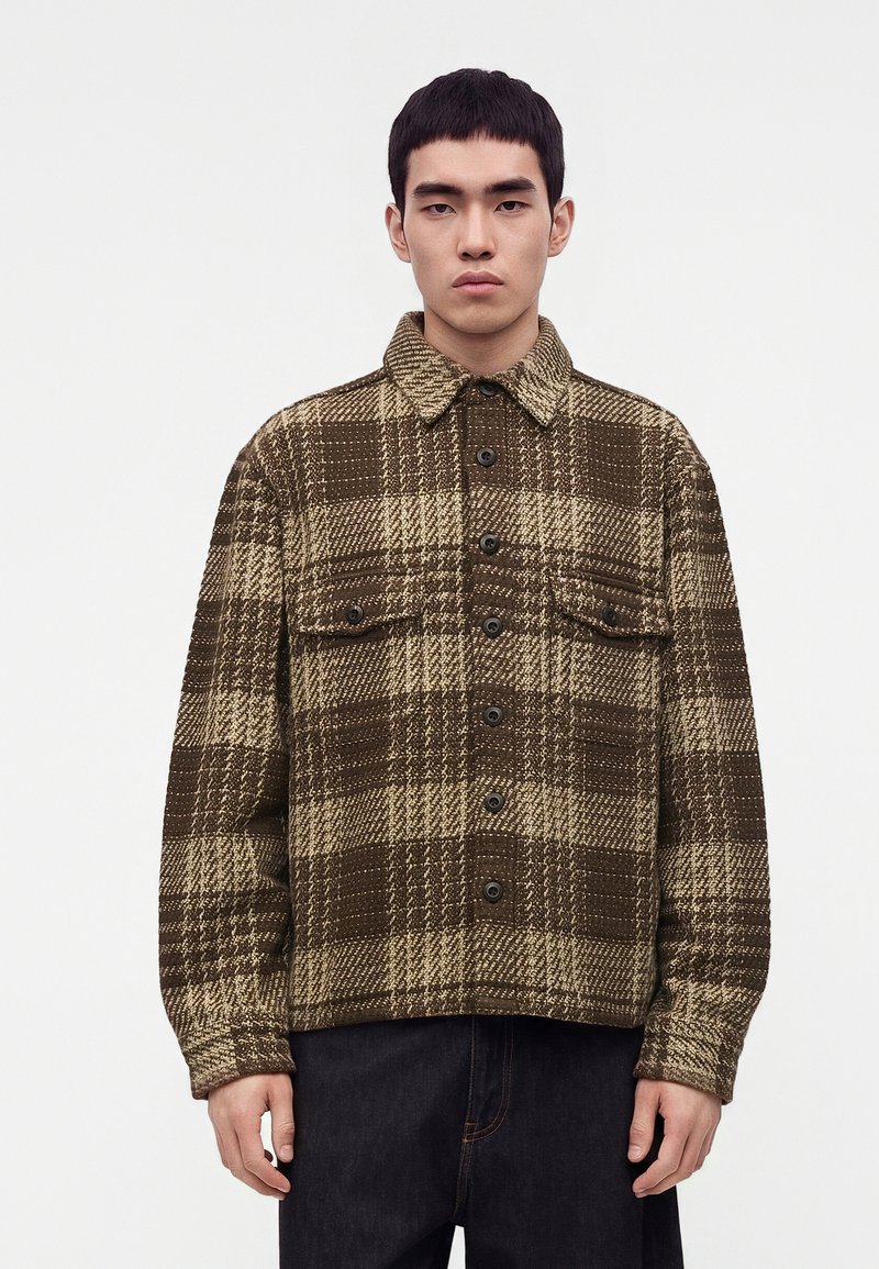 Jeune homme portant une veste à carreaux marron et beige boutonnée avec des poches poitrine, debout devant un fond blanc uni.
