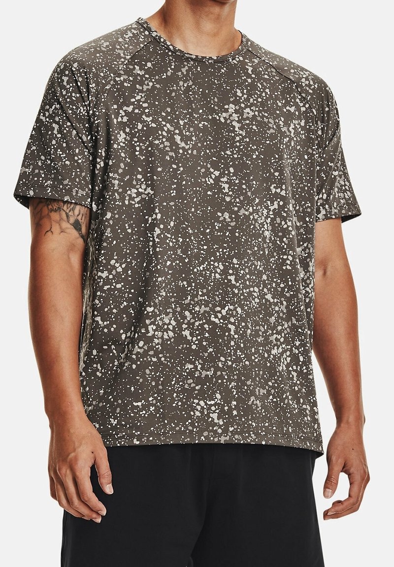 Homme portant un t-shirt marron à manches courtes avec un motif de projection de peinture blanche et un short noir, montrant un tatouage sur son bras gauche.