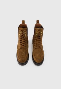 Botine din suede maro, cu șireturi, talpă neagră și urechițe aurii, văzute dintr-o parte, pe un fundal alb simplu.