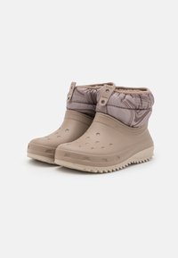 Bota de borracha bege com um upper texturizado, apresentando um topo almofadado e orifícios de ventilação. Sola robusta com padrão de tração. Detalhe de hardware ajustável.