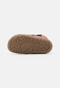 Apavi ar teksturētu brūnu gumijas zoli, kurai ir zigzaga raksts, reljefs logotips un izmēra indikators; siksnas uz rozā augšdaļas.