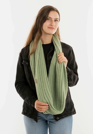 Groene gebreide infinity-sjaal met een gestructureerd ribpatroon, vastgehouden door een persoon in een zwarte jas en spijkerbroek tegen een witte achtergrond.