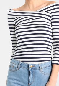 Granatowa i biała bluzka z długim rękawem w paski, z dekoltem bez ramion, wykonana z miękkiego materiału, zestawiona z jasnoniebieskimi spodniami z wysokim stanem.