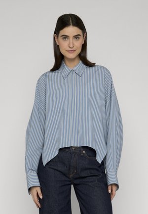 Junge Frau trägt ein locker sitzendes, hellblau-weiß gestreiftes Hemd mit Kragen, das teilweise in eine dunkelblaue Jeans gesteckt ist, und steht vor einem einfarbigen Hintergrund.