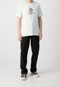 Světle zelené bavlněné tričko s krátkým rukávem, které má grafiku palmy a text. V kombinaci s černými rovnými kalhotami a plátěnými botami.