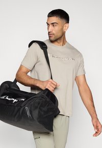 Γκρι μπλουζάκι με κεντημένο λογότυπο "Champion", συνδυασμένο με μαύρη τσάντα duffel με διακοσμητικά από δίχτυ και φερμουάρ, κρατημένη από έναν άνθρωπο.