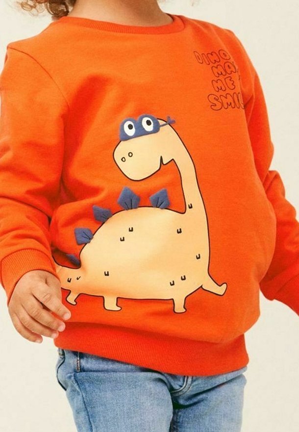 Enfant portant un sweat-shirt orange avec un dinosaure de dessin animé et le texte "Dino Make Me Smile", associé à un jean bleu.