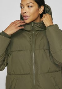 Olivgrön pufferjacka med hög krage och dragkedjestängning. Har horisontell sömnad och elastiska manschetter. Mjuk, vadderad textur.