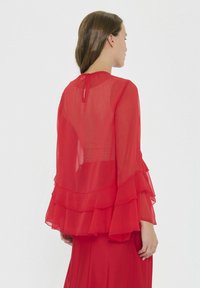 Rote, durchsichtige Bluse mit geschichteten Volants an den Ärmeln und dem Saum. Schlitzverschluss hinten; leichtes Gewebe mit glatter Textur.