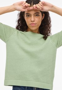 Γυναίκα με σγουρά μαλλιά που φοράει ένα ανοιχτό πράσινο υφασμάτινο πουλόβερ, σηκώνει τα χέρια κοντά στο μέτωπο μπροστά σε απλό λευκό φόντο.