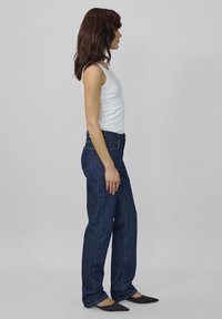 Žena s dlhými tmavými vlasmi po pleciach, oblečená v bielej bezramienkovej blúzke, tmavomodrých rovno strihaných džínsoch a čiernych špičatých topánkach, stojí bokom.