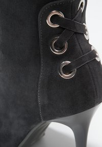Bota de ante negra hasta el tobillo con puntera afilada, tacón alto y diseño de cordones con ojales plateados y detalles entrelazados.
