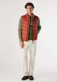Gilet imbottito color ruggine da uomo con colletto alto, abbinato a un maglione verde a trama, pantaloni bianchi e scarpe marrone scuro.